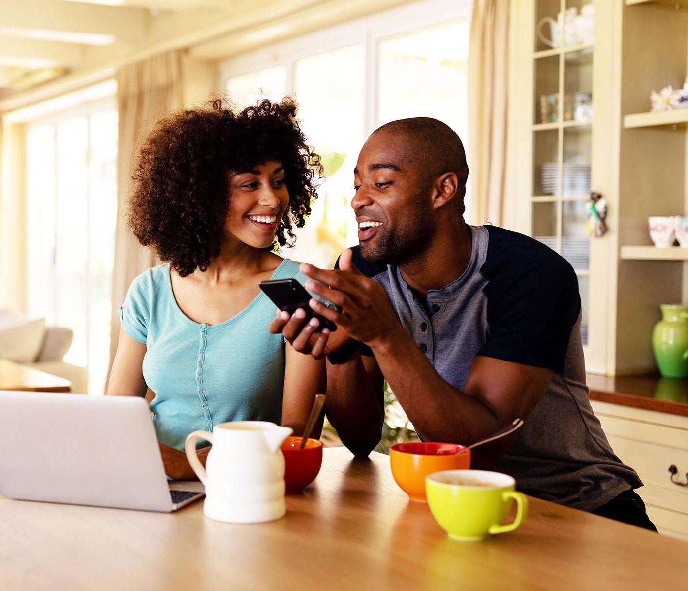 Couple looking at a smartphone with coffee and a laptop on the table.